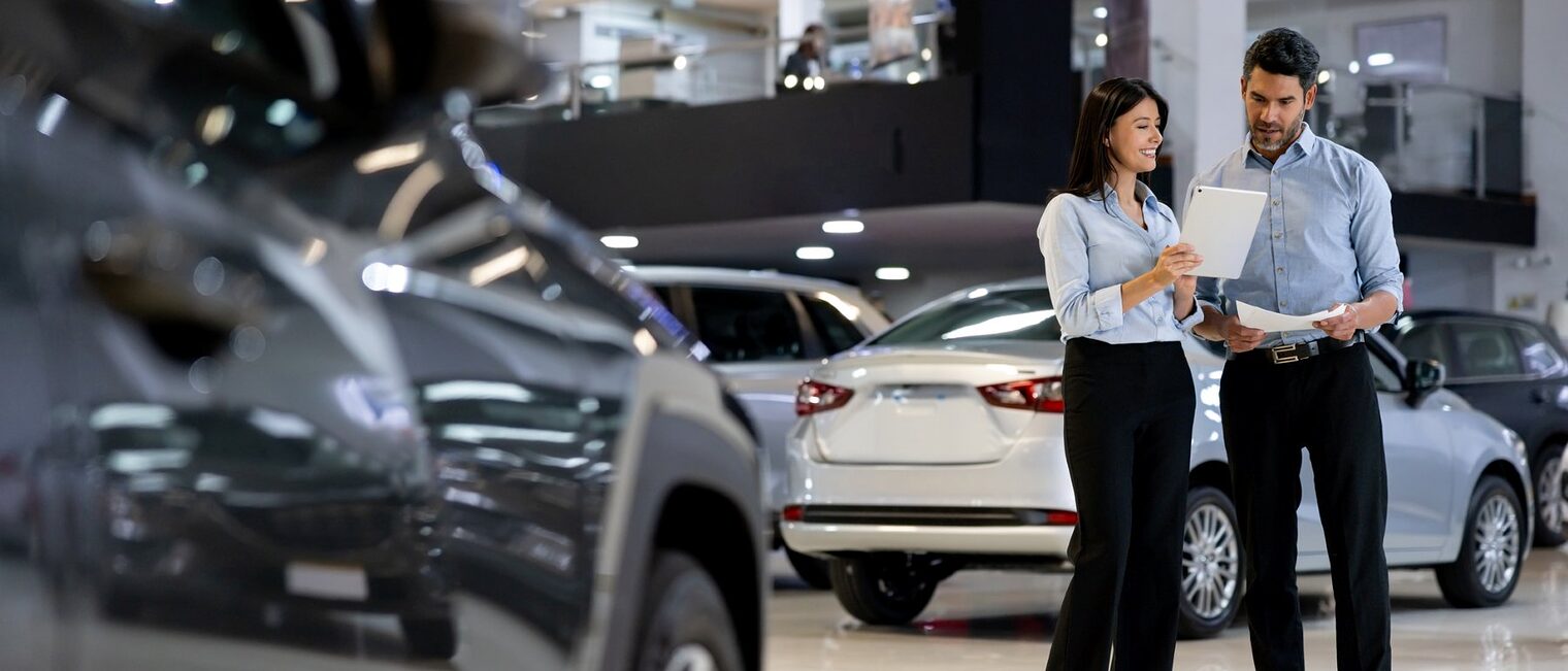 Eine Verkäuferin und ein Verkäufer in einem Autohaus, die gemeinsam Unterlagen durchgehen. Im Vordergrund ein Auto.