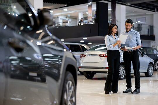 Eine Verkäuferin und ein Verkäufer in einem Autohaus, die gemeinsam Unterlagen durchgehen. Im Vordergrund ein Auto.