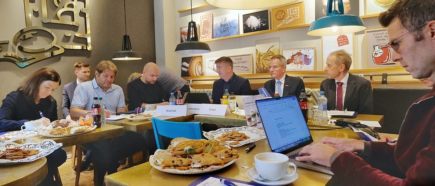 Teilnehmende der Pressekonferenz sitzen an Tischen in der Bäckerei. Es ist gemütlich eingedeckt mit Kuchen und Gebäck.