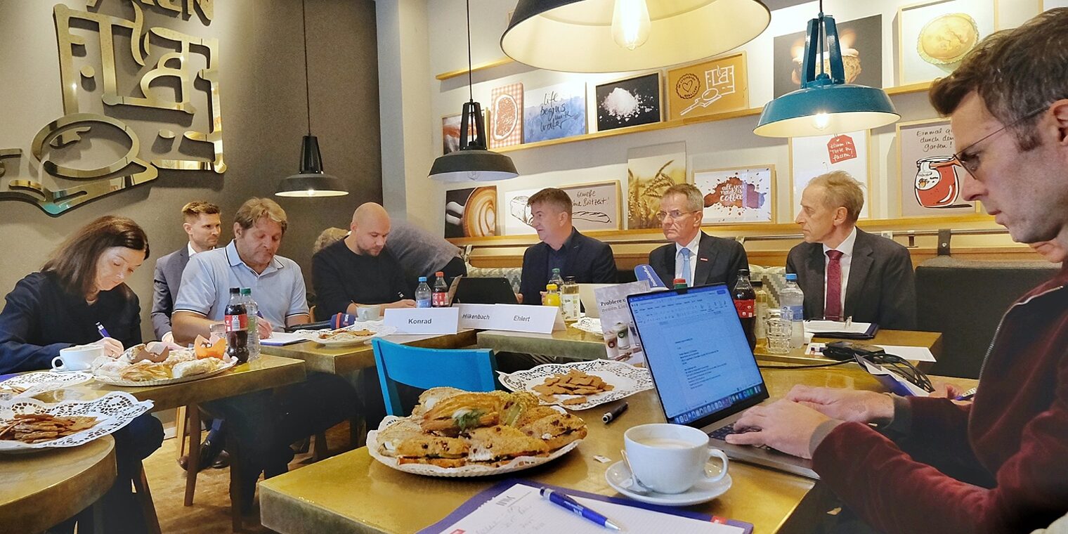 Teilnehmende der Pressekonferenz sitzen an Tischen in der Bäckerei. Es ist gemütlich eingedeckt mit Kuchen und Gebäck.
